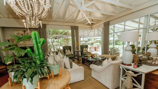 Amplia sala de estar con techo alto, ventanas grandes y varios muebles, incluyendo una mesa de madera, sofás blancos y una estatua decorativa de un perro.