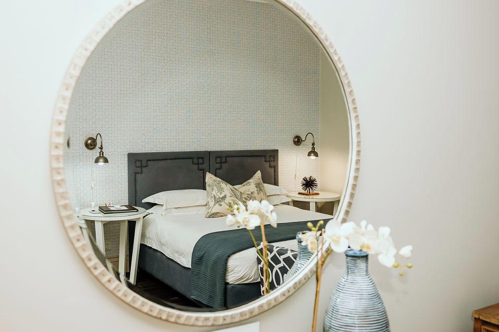 A bedroom with a round mirror, a bed with pillows, a bedside table with a lamp, and a vase with flowers.