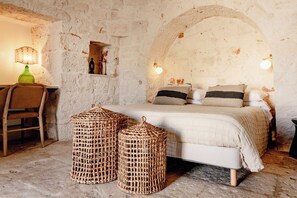 Ein rustikales Schlafzimmer mit Steinwand, einem Bett mit vielen Kissen, einem Sessel und zwei gewebten Hockern.