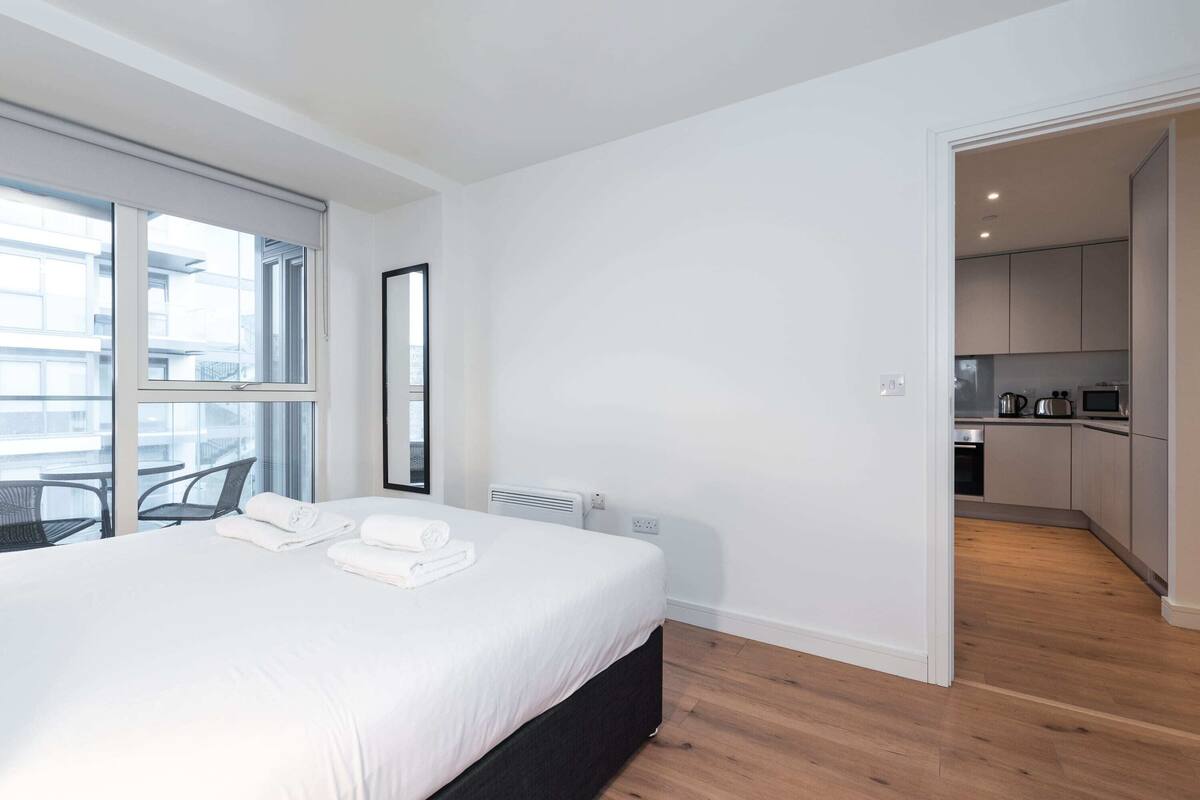 A modern hotel room with a large bed, a view through a sliding door to a balcony, and a kitchenette in the adjacent room.