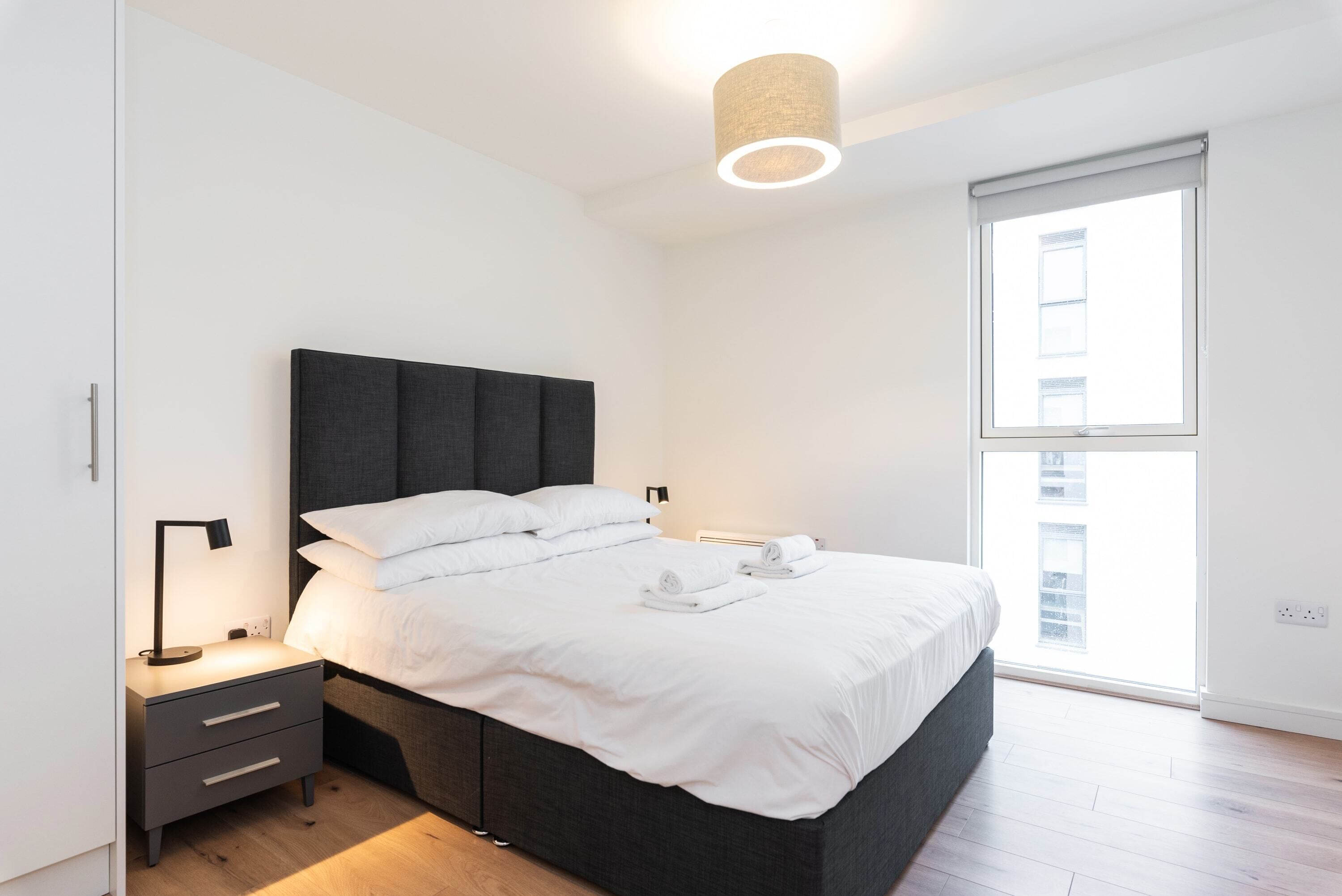 A modern bedroom with a large bed, a bedside table with a lamp, and a window with blinds.