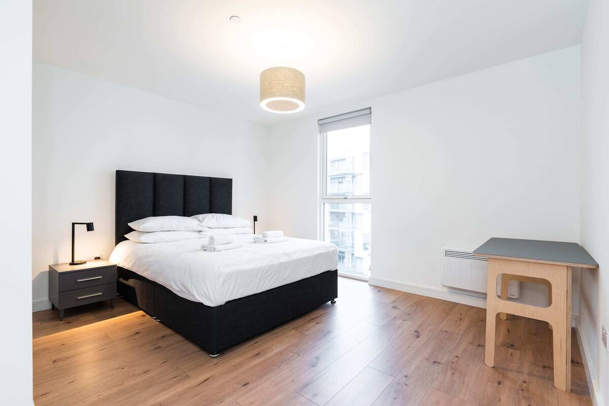 A modern bedroom with a bed, bedside table, lamp, and a small wooden desk.