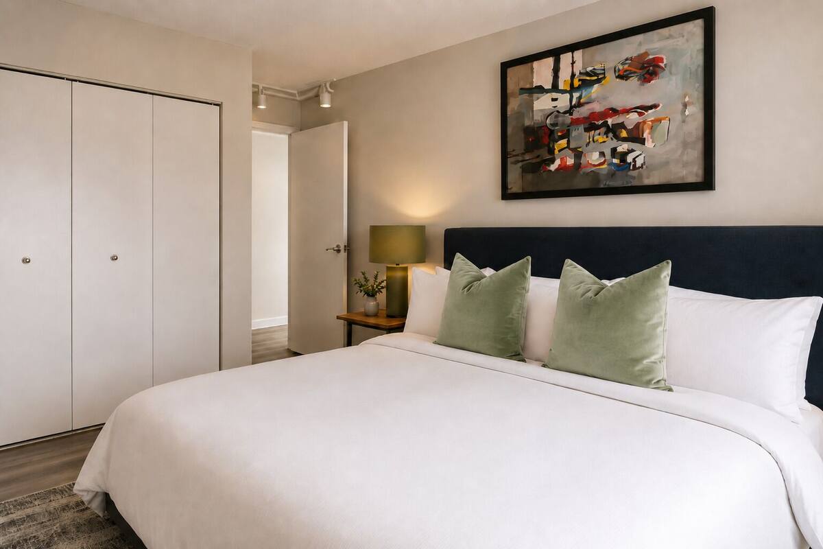 A hotel room with a large bed, a bedside table with a lamp, a painting on the wall, and a wardrobe.