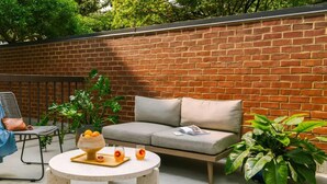 A patio area with a brick wall, a grey sofa, a round table with a bowl and glasses, and potted plants.