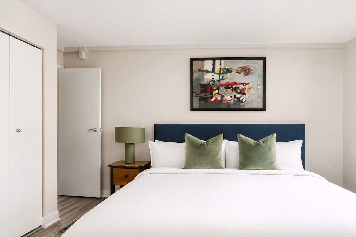 A bedroom with a bed, a nightstand with a lamp, a door, and a painting on the wall.
