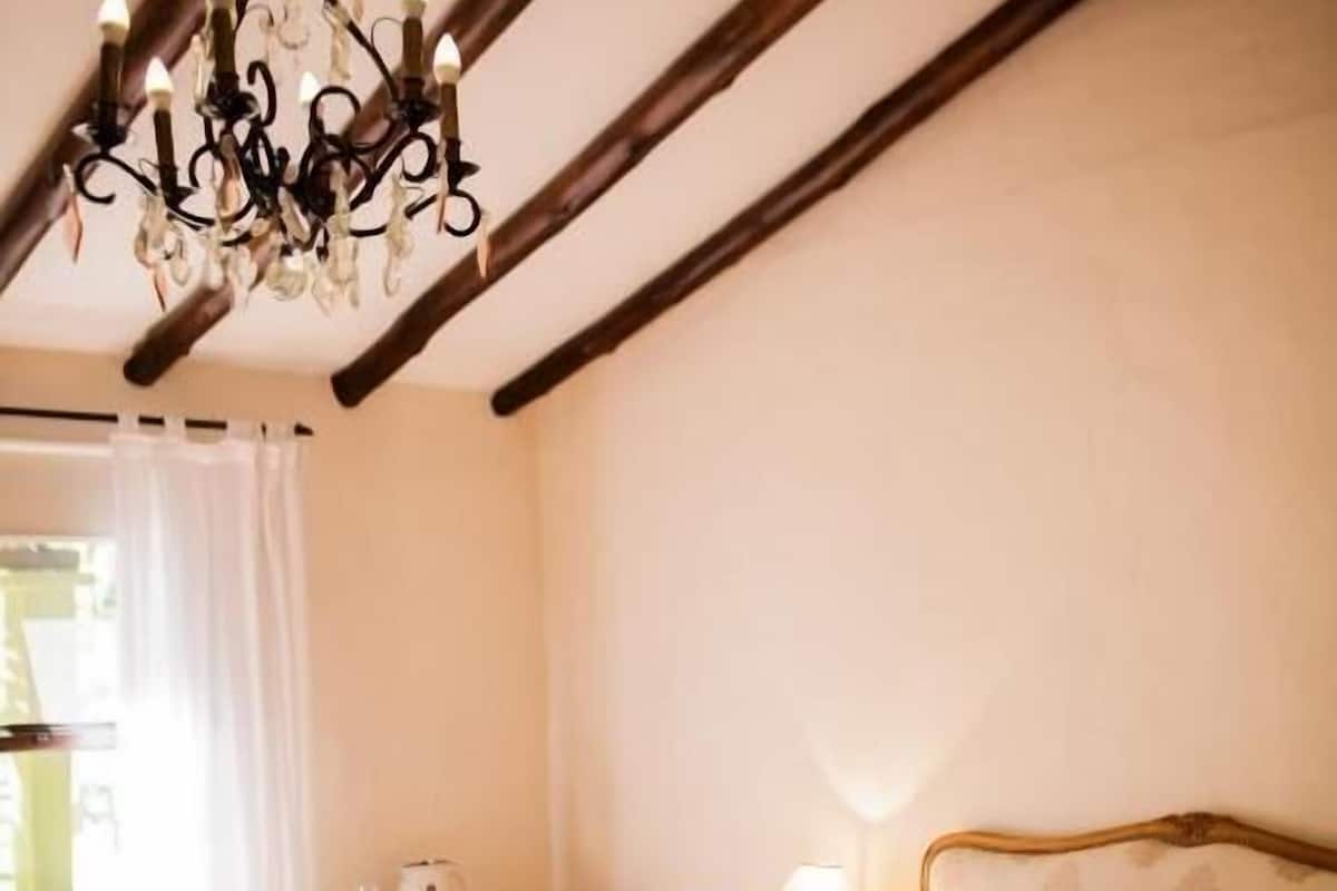 A bedroom with a wooden ceiling, a bed with a pink and white striped blanket, a bedside table with a lamp, and a window with curtains.