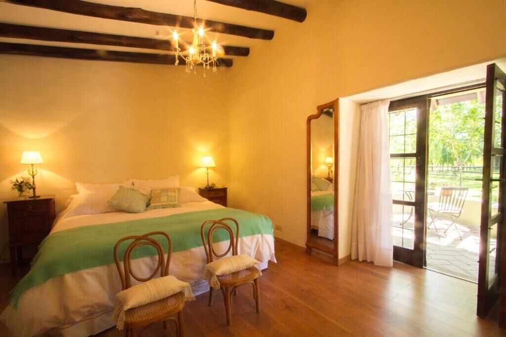Quarto com cama, duas mesas de cabeceira com luminárias, espelho, cadeira e porta de vidro corrediça para área externa.
