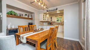 A dining area with a wooden table and chairs, a kitchen in the background, and a sign that reads 'It's Cook Something Somewhere.'