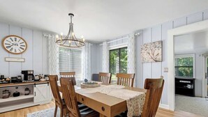 Comedor con mesa de madera, sillas, una araña, un reloj y una zona de cocina con armarios y electrodomésticos.