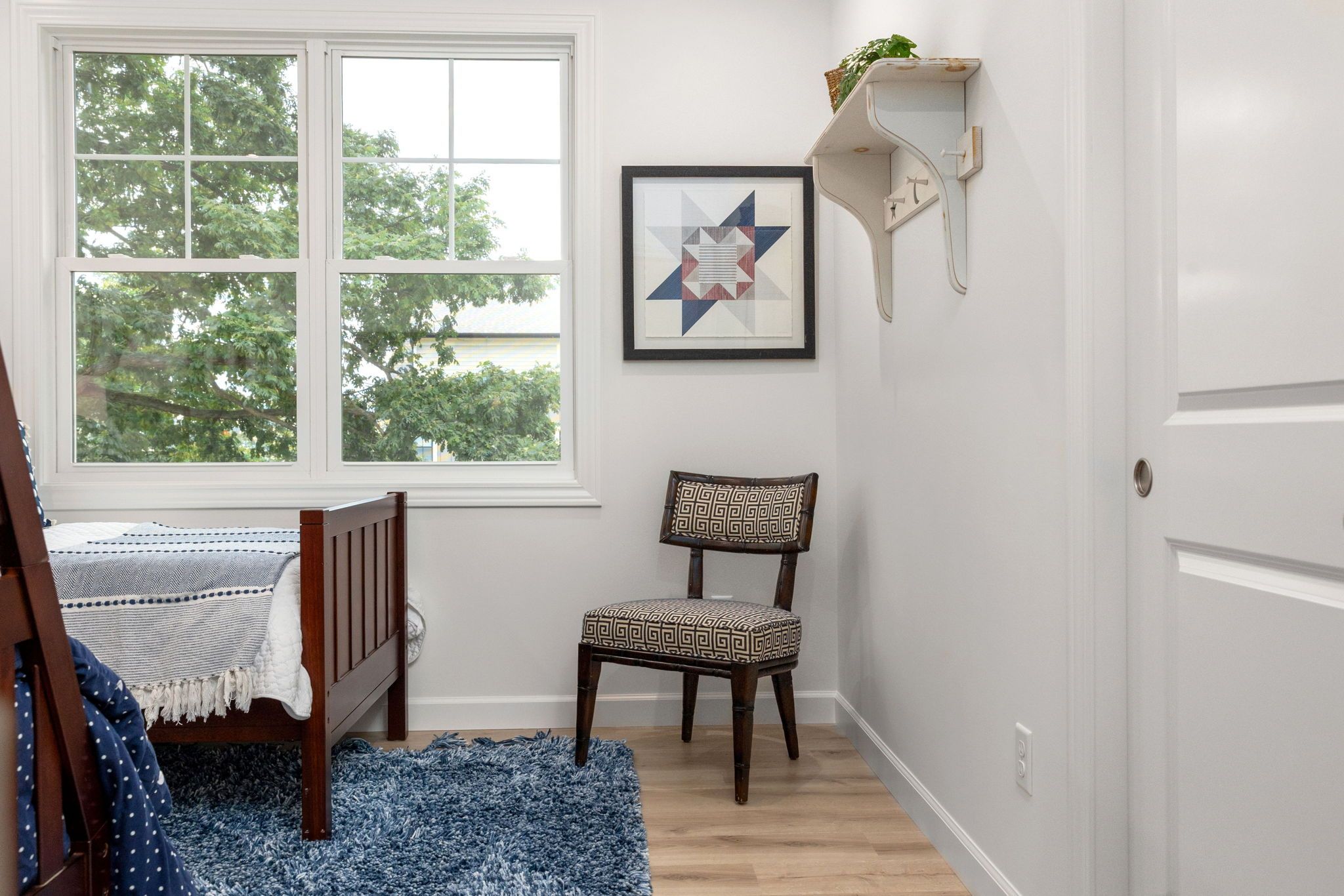 A room with a bed, a chair, a rug, a window, and a wall-mounted shelf with a framed picture.