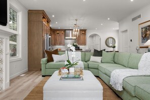 A modern living room with a green sectional sofa, a white coffee table, and a kitchen in the background. - Across From ND Stadium | Luxury 6B/5BA | Sleeps 14 (South Bend)