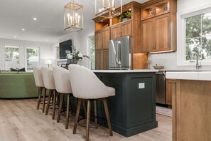 A modern kitchen with wooden cabinets, a stainless steel refrigerator, and a bar area with stools. - Across From ND Stadium | Luxury 6B/5BA | Sleeps 14 (South Bend)