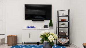 Ein Wohnzimmer mit einem an der Wand montierten Flachbildfernseher, einem weißen Schrank, einem Korbtisch, einem blau gemusterten Teppich und einer Vase mit Blumen.