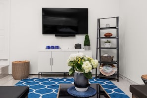 Ein Wohnzimmer mit einem an der Wand montierten Flachbildfernseher, einem weißen Schrank, einem Korbtisch, einem blau gemusterten Teppich und einer Vase mit Blumen.