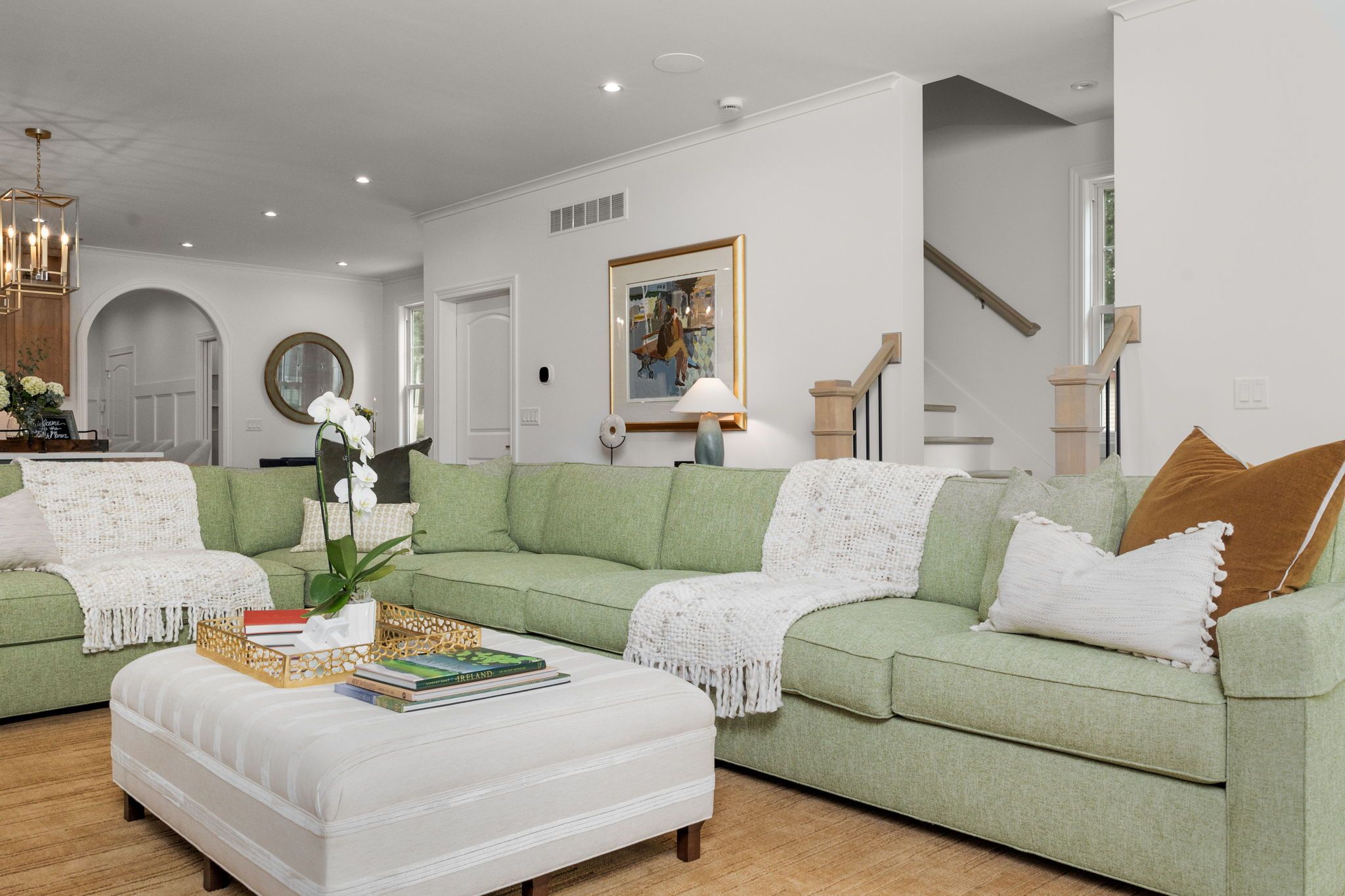Ein modernes Wohnzimmer mit einem grünen Sofa, einem weißen Hocker und einem Couchtisch mit Büchern. An der Wand hängt ein gerahmtes Bild und im Hintergrund ist eine Treppe zu sehen.