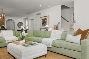 En moderne stue med en grøn sofa, hvid skammel og et sofabord med bøger. Et indrammet billede hænger på væggen, og en trappe ses i baggrunden.
