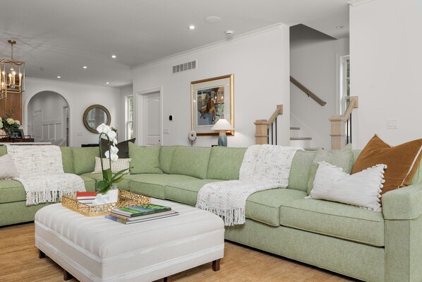 Een moderne woonkamer met een groene bank, een witte voetenbankje en een salontafel met boeken. Er hangt een lijst met een afbeelding aan de muur en een trap is in de achtergrond zichtbaar.