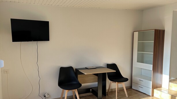 Een kamer met een wandtelevisie, een houten tafel met twee zwarte stoelen, een witte kast en een betegelde vloer.