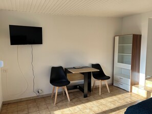 Ein Zimmer mit einem an der Wand befestigten Flachbildfernseher, einem Holztisch mit zwei schwarzen Stühlen, einem weißen Kleiderschrank und einem gefliesten Boden.
