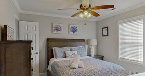 Ein Schlafzimmer mit einem Bett, einem Deckenventilator, einem Nachttisch, einer Lampe, einem Fenster mit Jalousien und Bildern an den Wänden.
