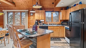 A wooden cabin kitchen with granite countertops, wooden cabinets, a black refrigerator, and a dining area with wooden chairs and a table. - Rustic Ridge Cabin 102 - Private Woodland Retreat near Devil's Lake State Park (Merrimac)