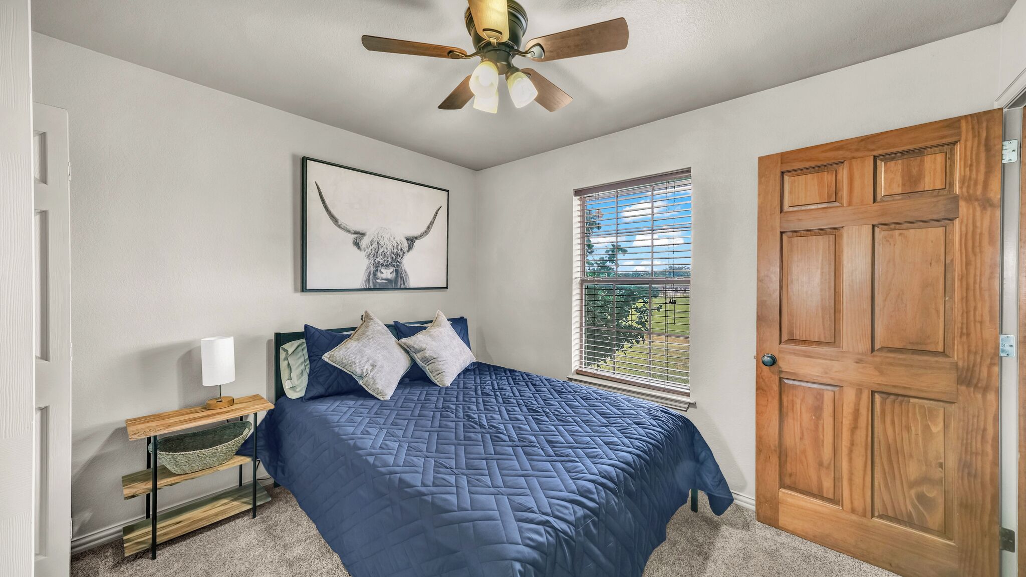 A bedroom with a bed, a ceiling fan, a window with blinds, and a wooden door.