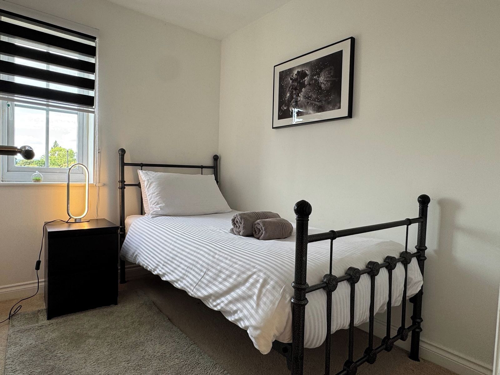 A single bed with a black metal frame, a bedside table with a lamp, and a framed picture on the wall.