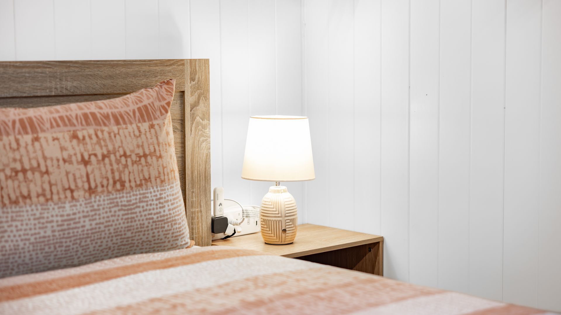 Un lit bien fait, agrémenté d’un coussin à motifs, d’une lampe de chevet et d’une prise électrique sur une table de chevet en bois.