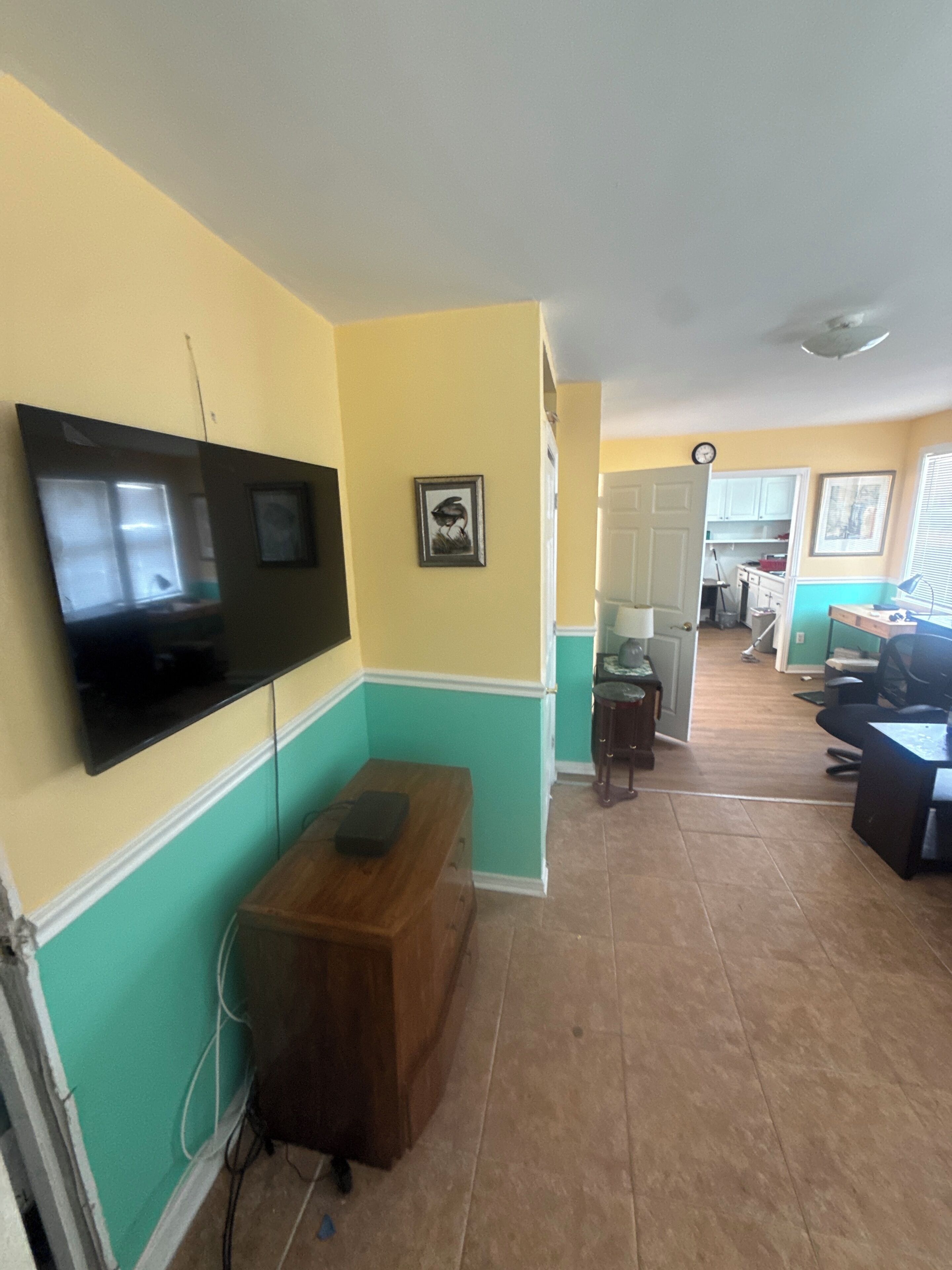 A room with a flat-screen TV mounted on the wall, a wooden bench, a door leading to another room, and a tiled floor.