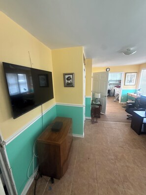 A room with a flat-screen TV mounted on the wall, a wooden bench, a door leading to another room, and a tiled floor.
