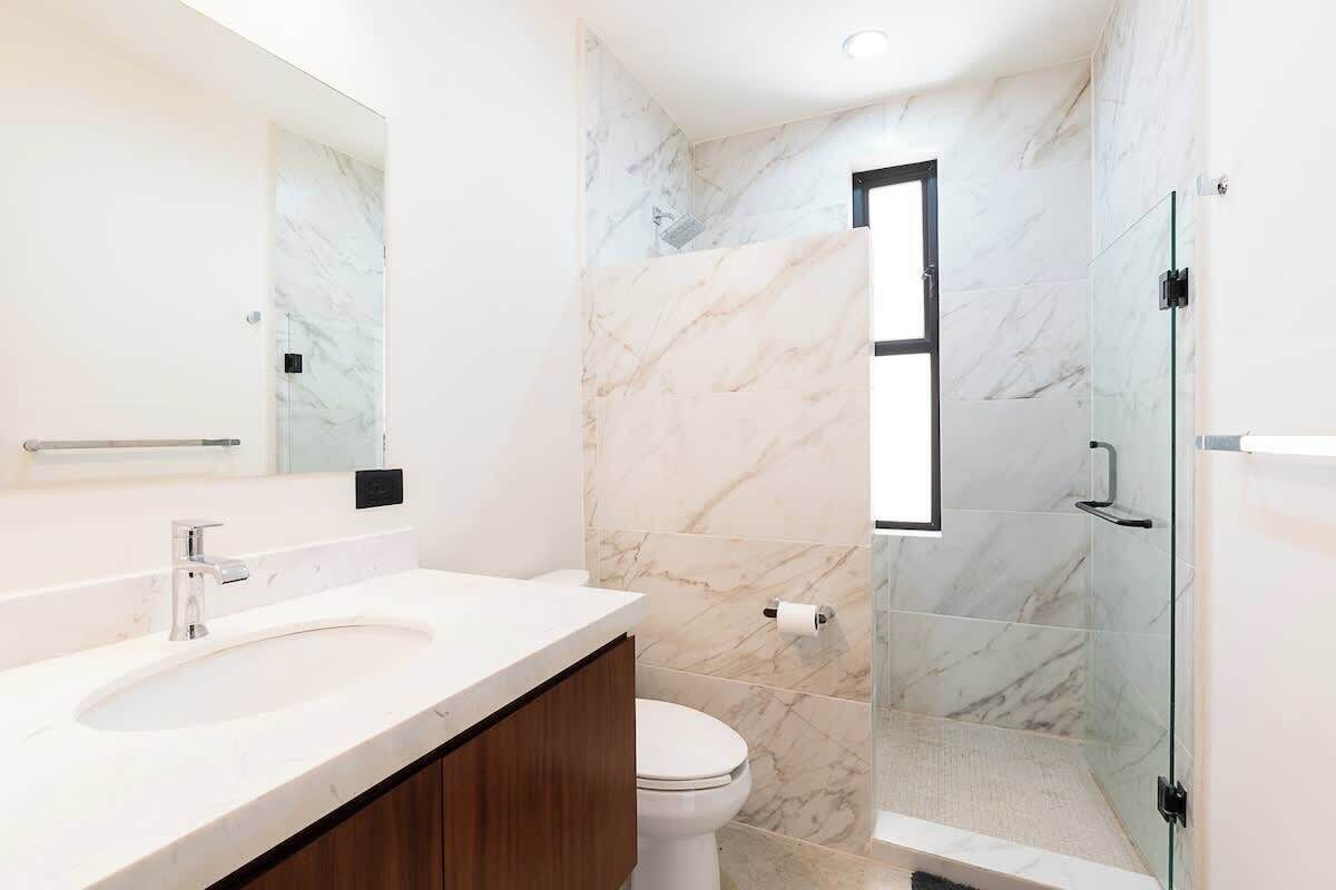 Un bany amb paret de marbre, un lavabo blanc, un vàter i una cabina de dutxa de vidre.