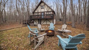 A cabin with a deck, surrounded by trees, and furnished with Adirondack chairs and a fire pit. - Cozy Cabin with Hot Tub, Game Room, Loft, Fire Pit (Pocono Lake)