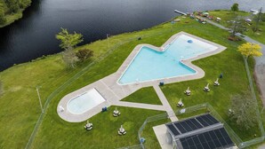 A swimming pool with a unique shape, surrounded by a fenced area with grass and a few lounge chairs. - Cozy Cabin with Hot Tub, Game Room, Loft, Fire Pit (Pocono Lake)