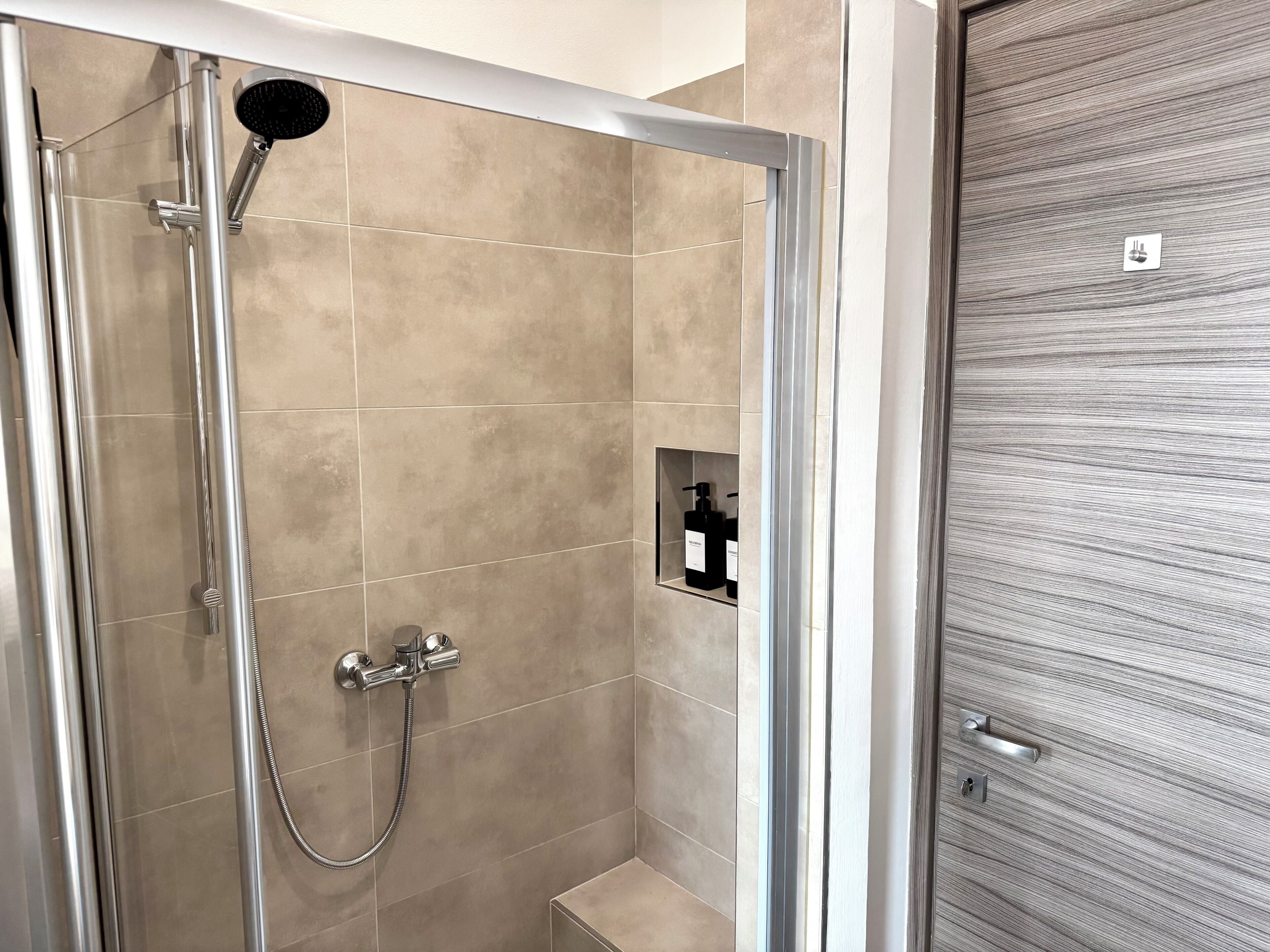 A shower with a handheld showerhead and a wall-mounted faucet, a small shelf with toiletries, and a door with a textured surface.