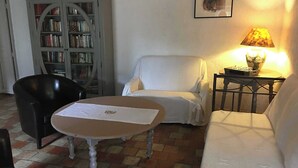 Una habitación con un sofá blanco, una silla negra, una mesa redonda con mantel blanco, una estantería y una lámpara sobre una mesita.