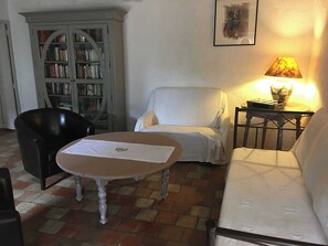 A room with a white sofa, a black chair, a round table with a white cloth, a bookshelf, and a lamp on a small table. - La Fromagerie - MONDION (MONDION)
