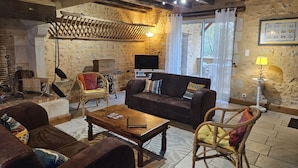 Ein rustikales Wohnzimmer mit Steinwand, Holzbalken, Kamin, einer Couch, einem Couchtisch und Korbmöbeln.