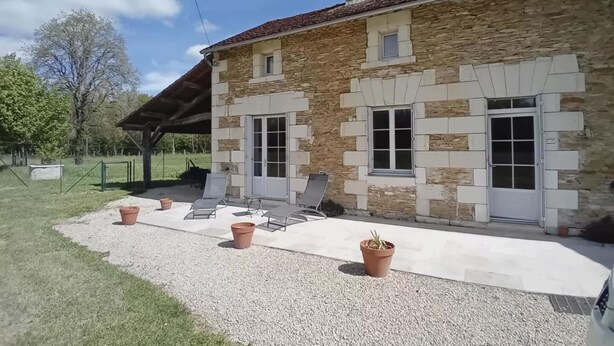 Un edificio in pietra con tetto spiovente, un portico coperto con sedie e un vialetto sterrato.
