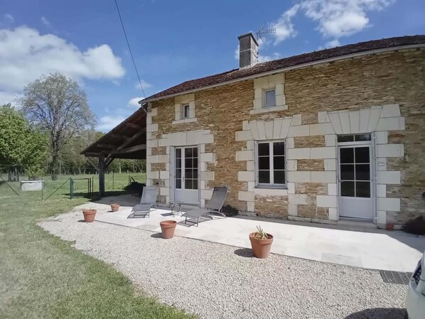 Ein Gebäude aus Stein mit einem geneigten Dach, einer überdachten Terrasse mit Stühlen und einer Zuwegung mit Kies.