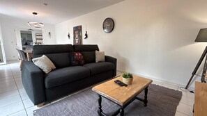A modern living room with a black sofa, a wooden coffee table, a clock on the wall, and a kitchen in the background. - Le Gîte de Bré - Near Futuroscope (CHENECHE)