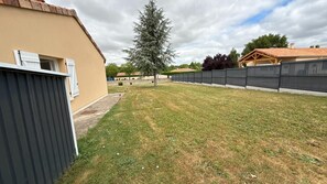 A residential backyard with a lawn, a tall evergreen tree, a black fence, and a beige building with a window. - Le Gîte de Bré - Near Futuroscope (CHENECHE)