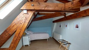 A room with a sloped ceiling, wooden beams, a bed with a white nightstand, a radiator, and framed pictures on the wall.
