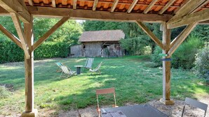 A wooden gazebo with a view of a backyard, including a lawn, two striped chairs, and a small table.