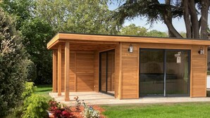 A modern wooden building with a covered patio area, large sliding glass doors, and a well-maintained garden with various plants and flowers.