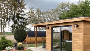 A wooden structure with a covered patio, a glass door, and a small garden with a hedge and potted plants.