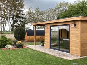 Una construcción de madera con un porche cubierto, una puerta de cristal y un pequeño jardín con seto y plantas en macetas.