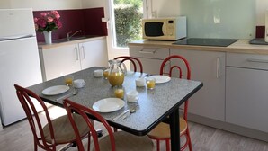 Een kleine keuken met een koelkast, magnetron en kookplaat, een eettafel met stoelen, en een raam met uitzicht op groen.