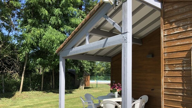 Een overdekt terras met een tafel en stoelen, omgeven door een houten hek en bomen.