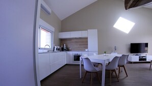 Et moderne køkken med hvide skabe, et spiseområde med et hvidt bord og stole, samt et fladskærms-tv monteret på væggen.