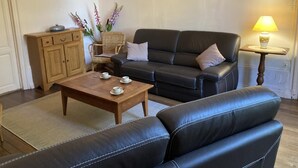 A living room with a sofa, a wooden coffee table, a wicker chair, a cabinet, and a lamp. - Le Petit Gîte du Theil Au Servant (LA CHAPELLE-VIVIERS)